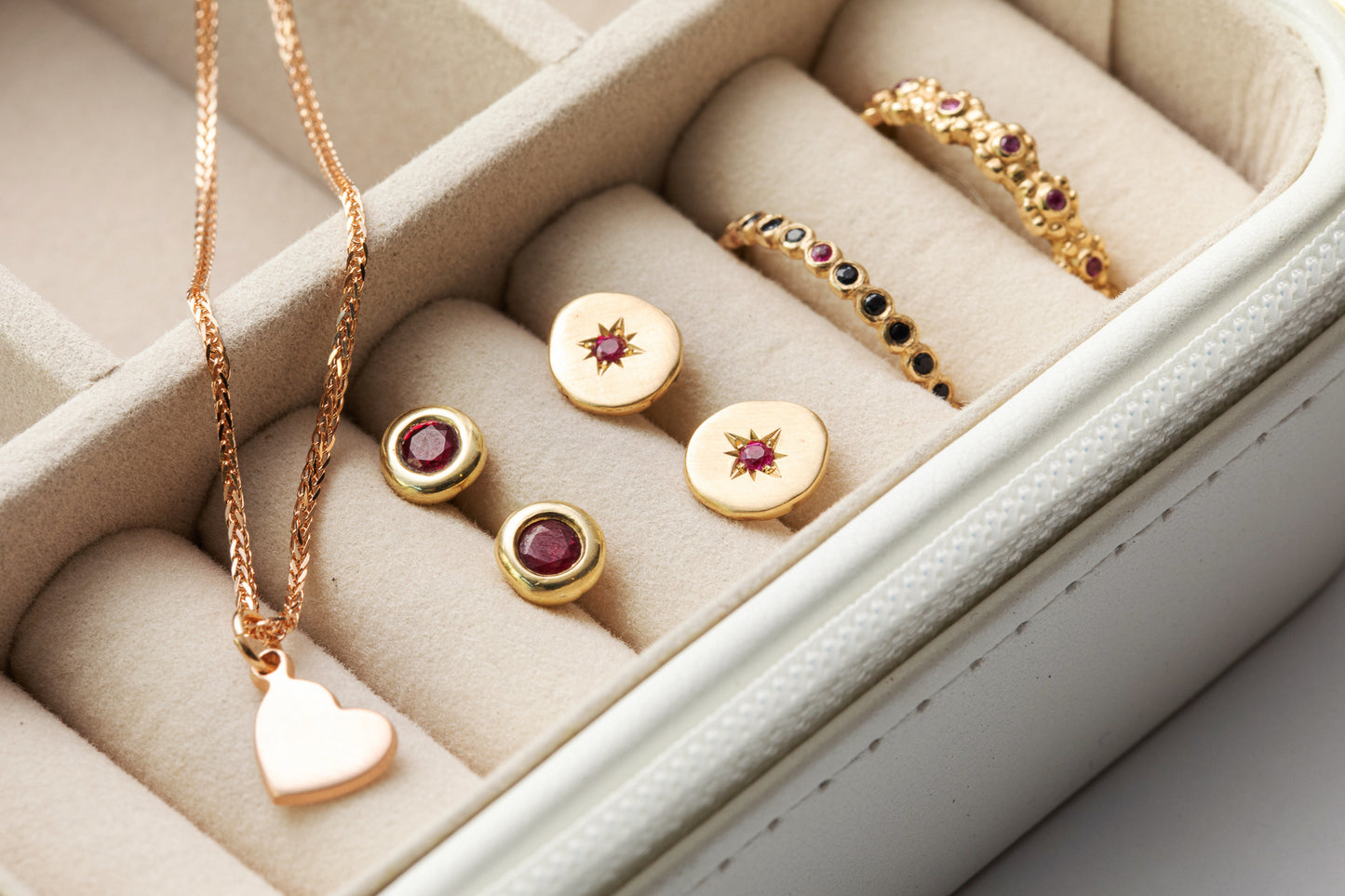 קופסת תכשיטים כוללת שרשרת תליון לב, שני זוגות עגילים עגולים עם אבני חן אדומות מרהיבות ושתי טבעות זהב מעוטרות באבני רובי קטנות מקולקציית טבעת זהב פרחים משובצת - רובי של Hydra Jewelry Studio, הכל בתא בצבע שמנת.
