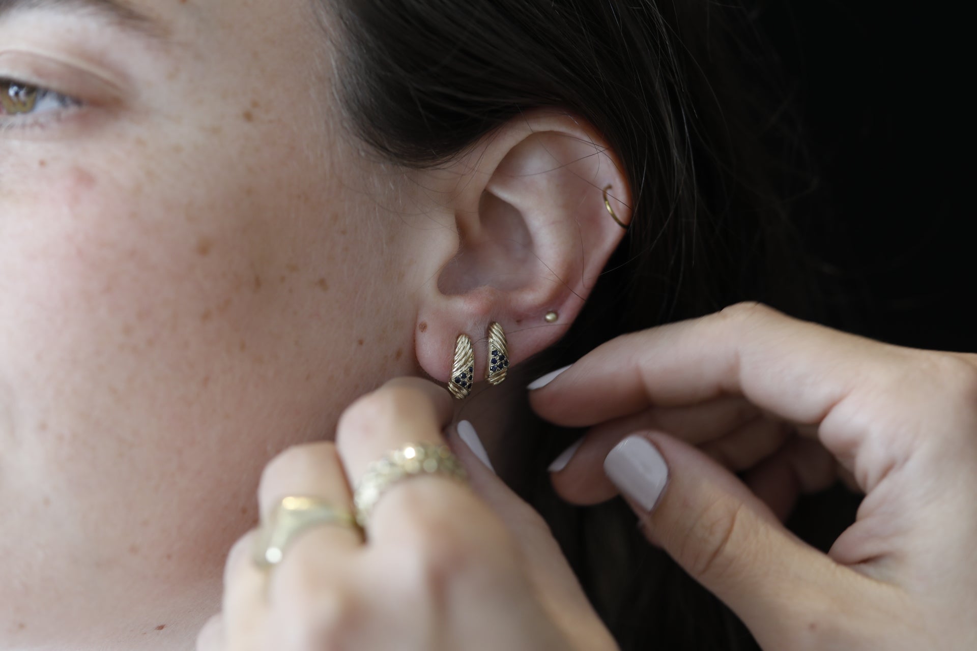 אוזן של אדם מעוטרת בעגיל זהב וינטאג׳- מורן - ספיר של סטודיו Hydra Jewelry, עגיל חישוק זהב מעוטר משופר באבני ספיר. יד נוספת, עם לק בגוון בהיר, מתאימה בקפידה את העגיל על רקע כהה שמדגיש יפה את פרטי הוינטג' הללו.