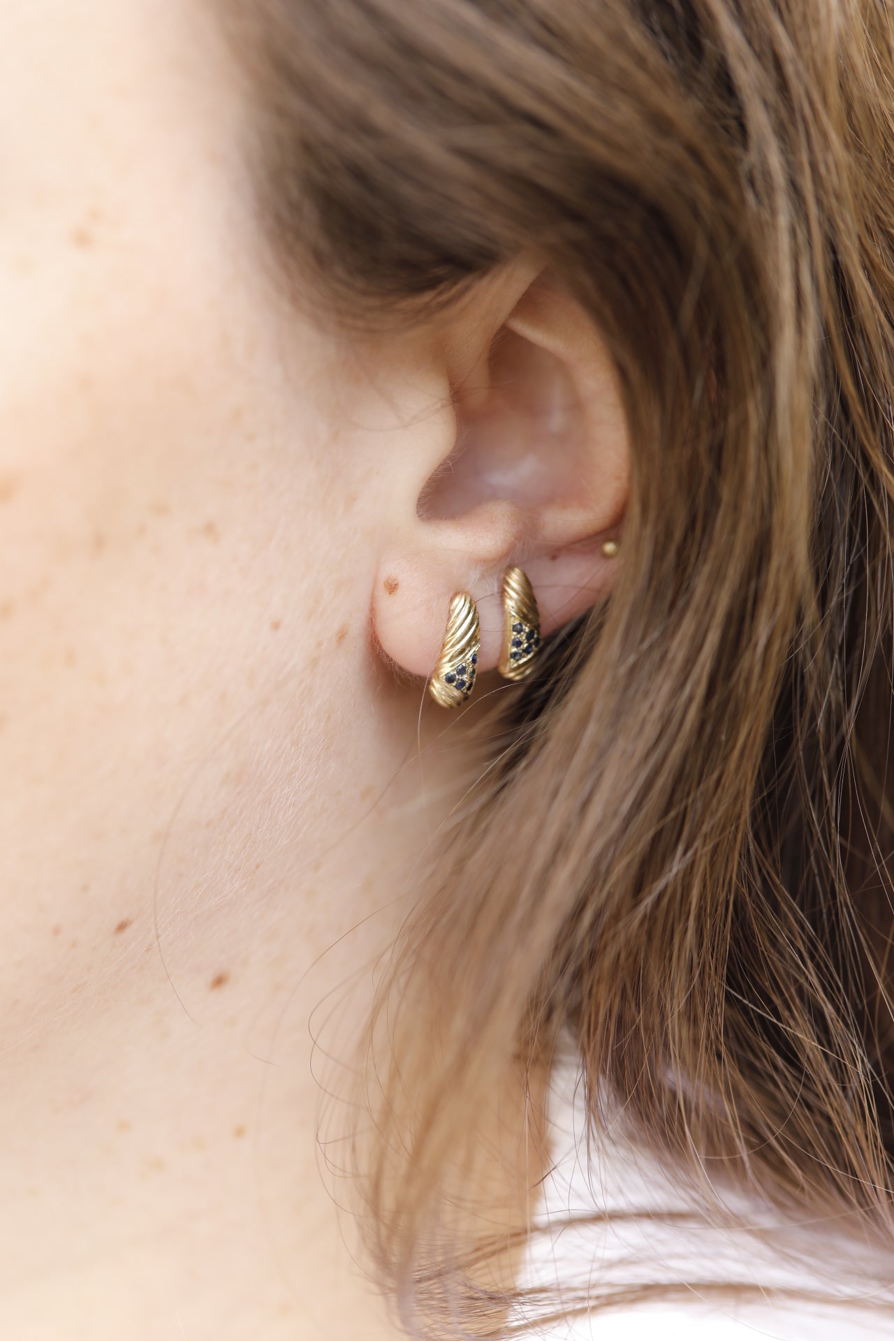 תקריב של אוזן מעוטרת בשני עגיל זהב וינטאג׳- מורן - ספיר מאת Hydra Jewelry Studio, הכולל חישוקי זהב עם עיצובים דמויי כנפיים ואבני ספיר כהות, מוסתרים חלקית בשיער חום.