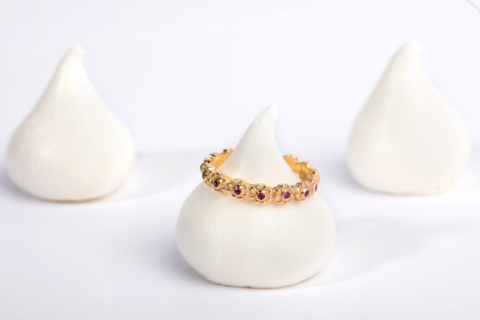 טבעת זהב פרחים משובצת - רובי מאת Hydra Jewelry Studio ממוקם באלגנטיות על גבי ממתקים דמויי מרנג עם שני פינוקים דומים מטושטשים בעדינות ברקע, כולם על רקע לבן ופשוט.
