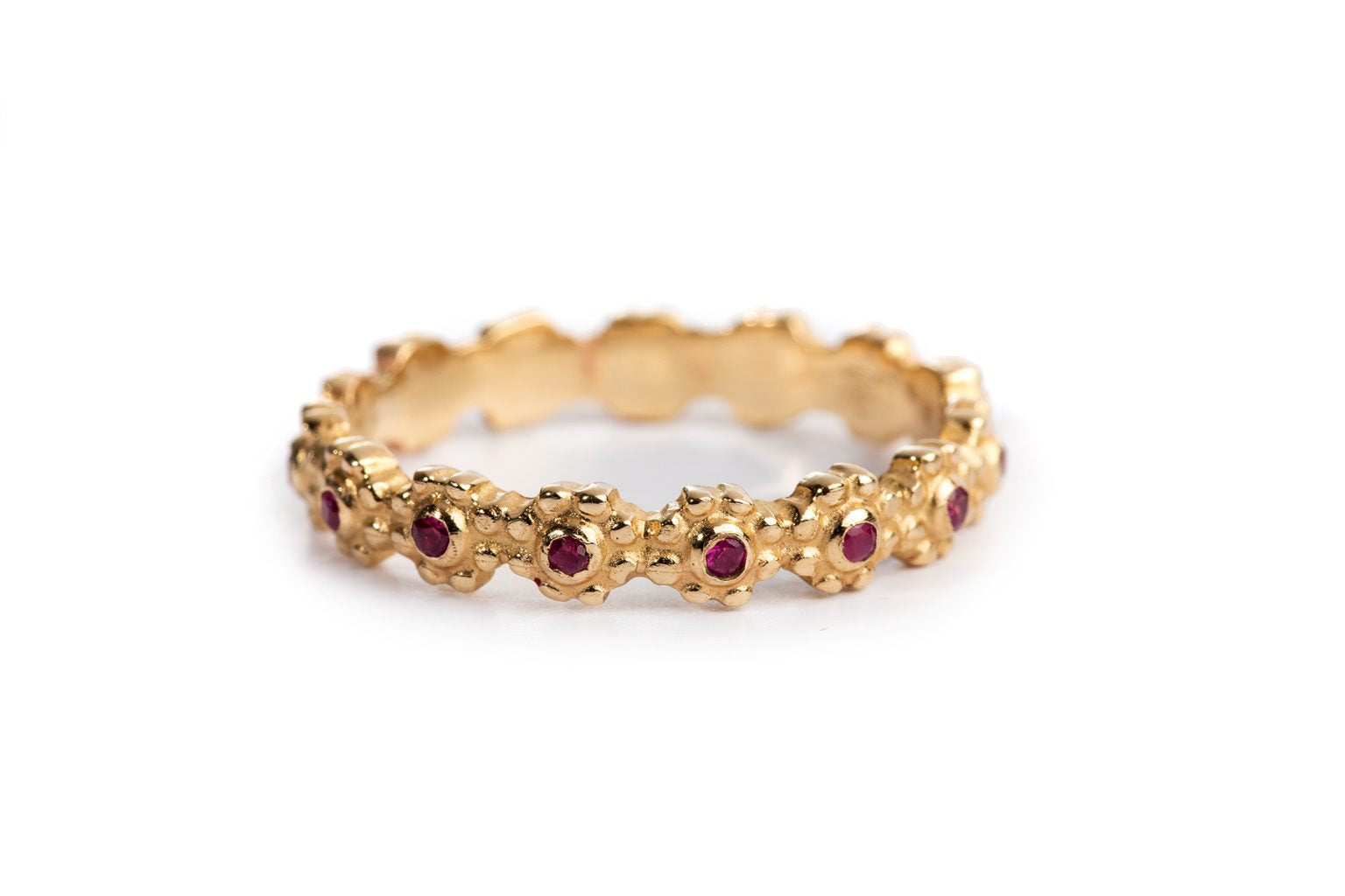 טבעת זהב פרחים משובצת - רובי של Hydra Jewelry Studio היא טבעת זהב עם רצועה בעלת מרקם, הכוללת אבני חן אדומות קטנות המזכירות אבן רובי, מוטבעות באופן שווה סביב הרקע הלבן שלה.