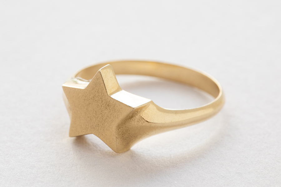 gold signet ring with a star shaped signet. made of 14 karat gold. hand made with a matte finish its shape is widest on top with the star reaching 11mm and becomes narrow at the back of the band reaching 2.5mm