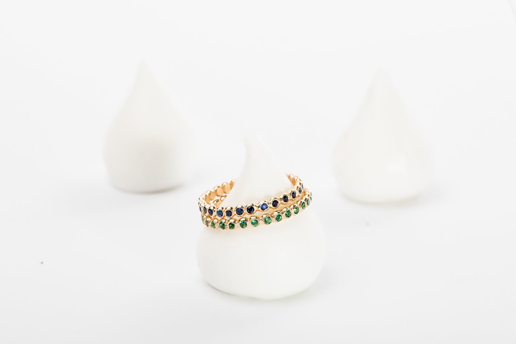 שתי טבעות זהב עם אבני חן אמרלד (טבעת עיגולי זהב - סופר מיני - אמרלד) של Hydra Jewelry Studio מוצגות על מעמד חלק, לבן, בצורת חרוט, כשברקע שני קונוסים מטושטשים דומים על משטח לבן.