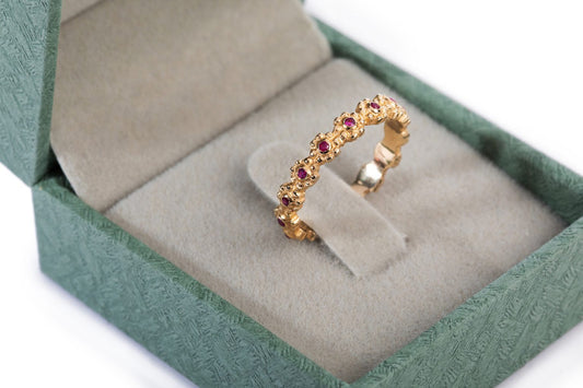 קופסת התכשיטים הפתוחה במרקם ירוק חושפת טבעת זהב פרחים משובצת - רובי מסטודיו Hydra Jewelry, הממוקמת באלגנטיות על פנים קטיפה אפור. העיצוב המורכב שלו מציג דוגמה המזכירה פרחי זהב, ויוצר אפקט קסום.