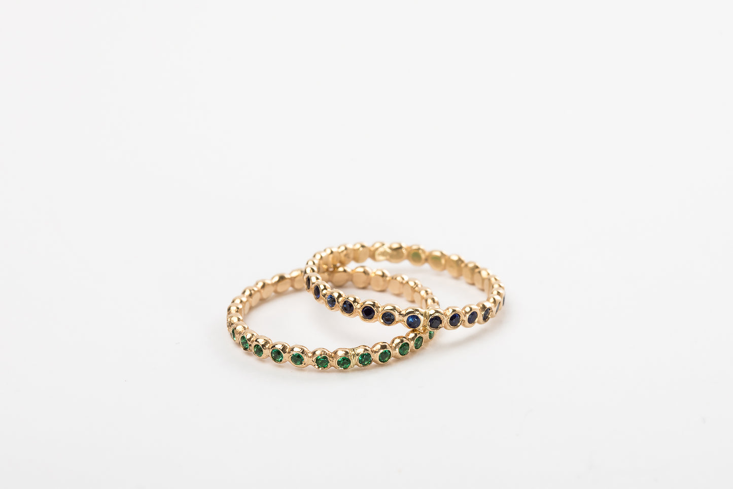 שתי טבעות זהב עדינות מסטודיו Hydra Jewelry - האחת, "טבעת עיגולי זהב - סופר מיני - אמרלד", עם אבני אמרלד ירוקות, והשנייה משובצת אבנים כחולות כהות - חופפות על רקע לבן רגיל.