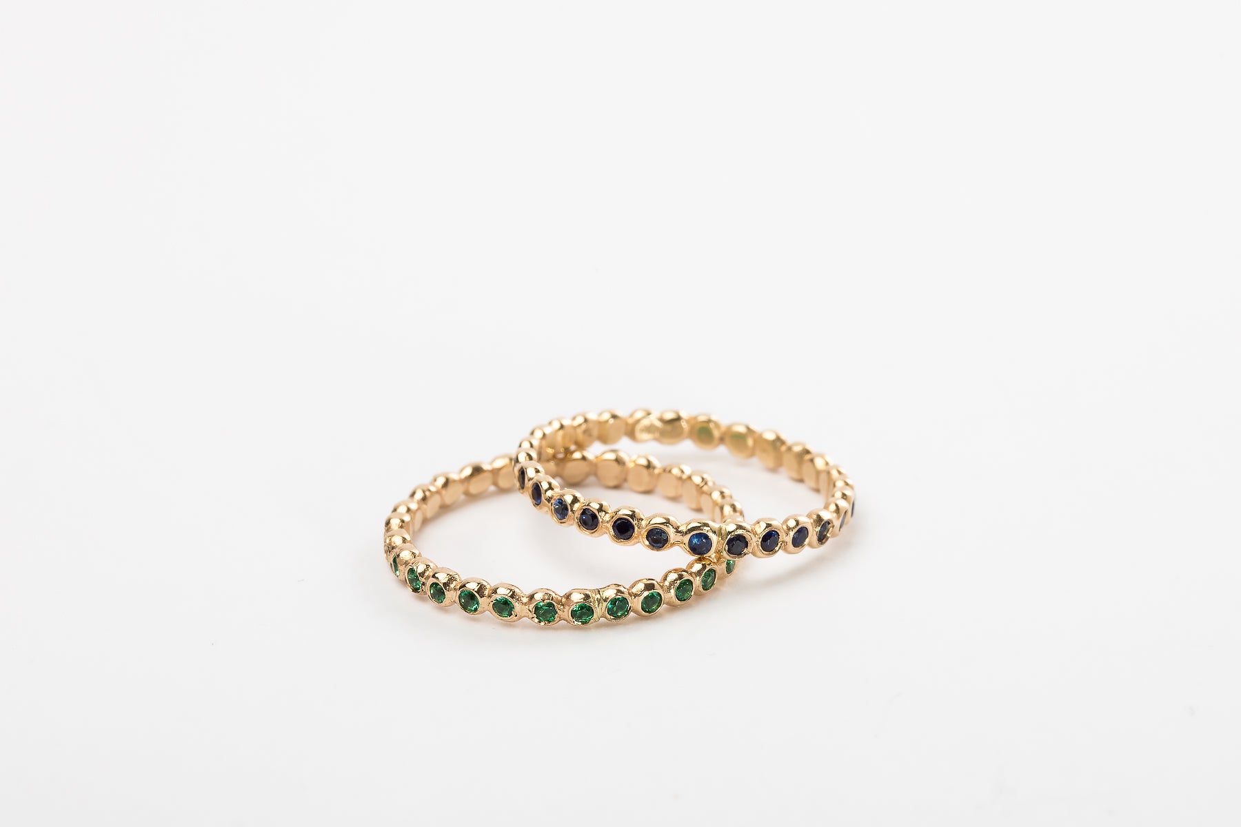 שתי טבעות זהב עדינות מסטודיו Hydra Jewelry - האחת, "טבעת עיגולי זהב - סופר מיני - אמרלד", עם אבני אמרלד ירוקות, והשנייה משובצת אבנים כחולות כהות - חופפות על רקע לבן רגיל.