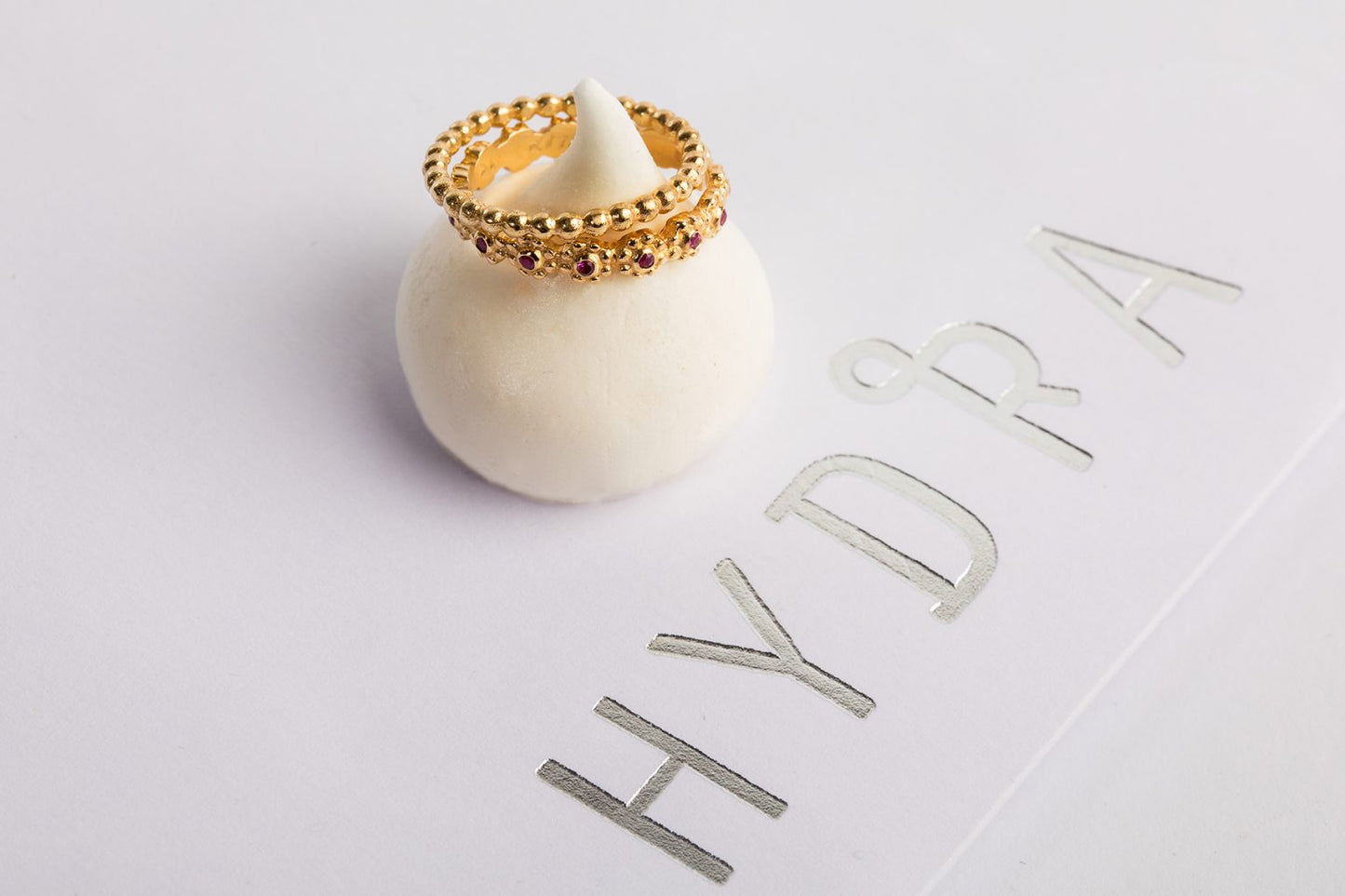 טבעת זהב עם אבני חן ורודות, בדומה לטבעת זהב פרחים משובצת - רובי, מוצגת על מעמד כדורי לבן. "HYDRA" מ-Hydra Jewelry Studio מוטבעת בכסף על המשטח הלבן שלצידה.