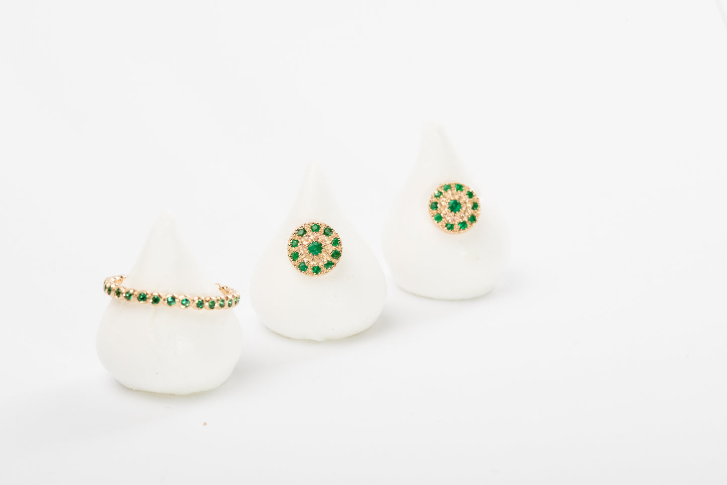 שלושה קונוסים לבנים מציגים יצירות של Hydra Jewelry Studio: משמאל טבעת טבעת עיגולי זהב - סופר מיני - אמרלד; במרכז ובימין, עגילים עגולים עם אבני אמרלד וחרוזי זהב עדינים - הכל על רקע לבן רגיל.