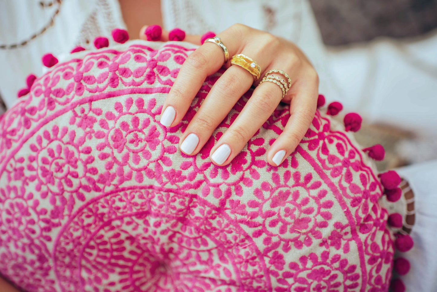 תקריב מציגה יד עם לק לבן מעוטרת בטבעות זהב, כולל טבעת זהב פרחים משובצת - רובי של סטודיו Hydra Jewelry, מונחת על כרית עגולה בדוגמת ורוד ולבן הכוללת רקמה פרחונית מורכבת ופונפונים.