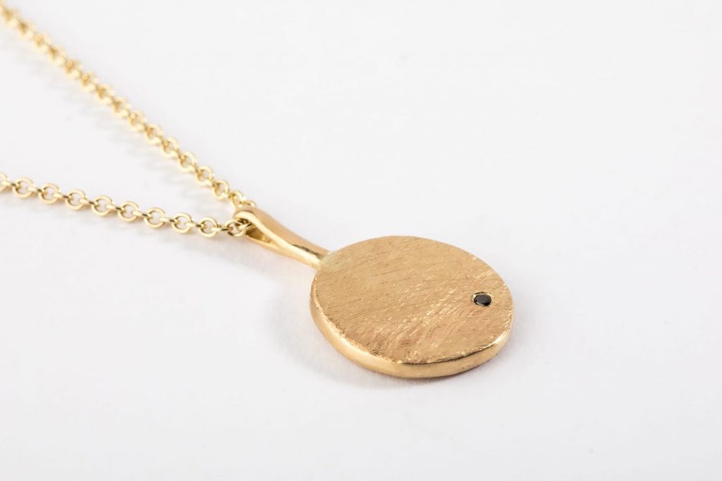 שרשרת זהב עם תליון בצורת מטבע זהב שעליו משובצת אבן יהלום שחור עדינה. המטבע מחובר עם וו זהב. 