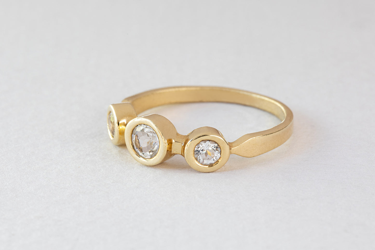 טבעת זהב אביב - טופז לבן של Hydra Jewelry Studio היא טבעת זהב הכוללת שלושה יהלומים עגולים בשוליים עגולים, משובצים באלגנטיות על משטח אפור בהיר, המזכיר את טבעת אביב.