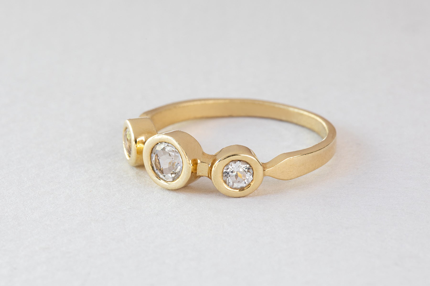 טבעת זהב אביב - טופז לבן של Hydra Jewelry Studio היא טבעת זהב הכוללת שלושה יהלומים עגולים בשוליים עגולים, משובצים באלגנטיות על משטח אפור בהיר, המזכיר את טבעת אביב.