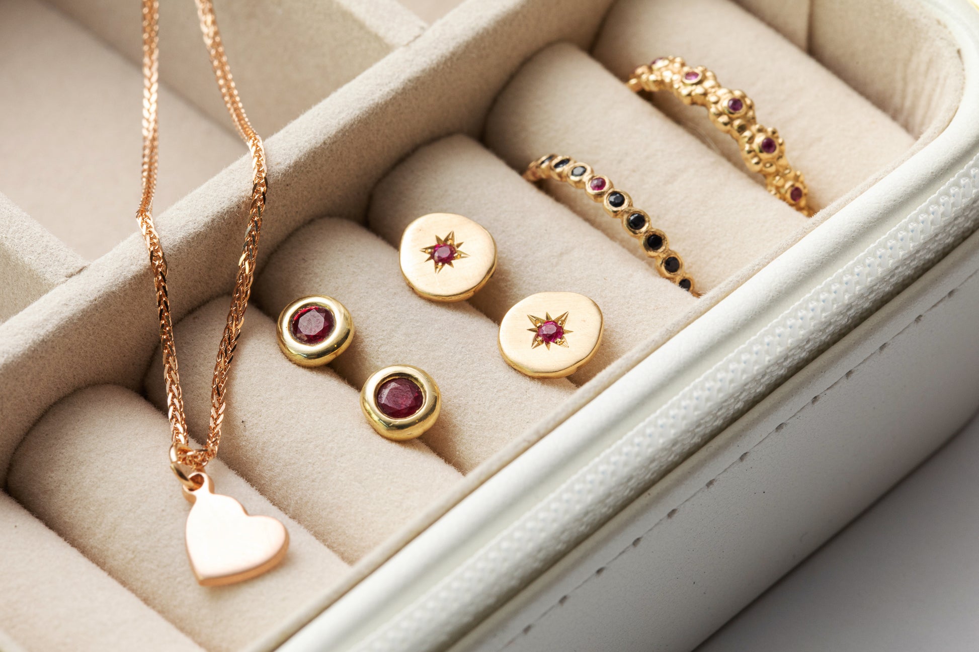 קופסת תכשיטים כוללת שרשרת תליון לב, שני זוגות עגילים עגולים עם אבני חן אדומות מרהיבות ושתי טבעות זהב מעוטרות באבני רובי קטנות מקולקציית טבעת זהב פרחים משובצת - רובי של Hydra Jewelry Studio, הכל בתא בצבע שמנת.